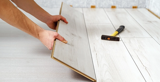 A person places a light-colored laminate flooring plank while a hammer rests on the floor nearby.